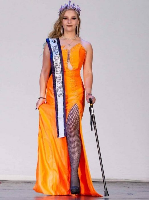 Contributor wearing an orange gown with sash and crow and using a cane to walk down the runway