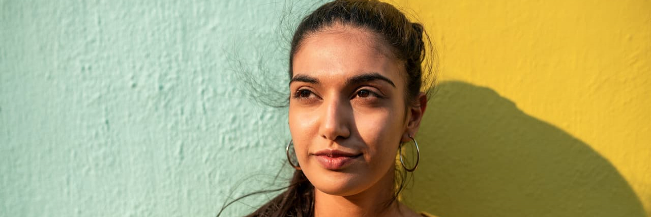 What I Wish I Could Tell My Younger Self About Life With Schizophrenia Portrait of Indian woman looking away while leaning on light blue/ yellow wall