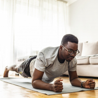 Confident muscled young man wearing sport wear and doing plank position while exercising on the floor in home interior.