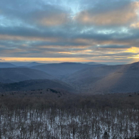 Dark mountains at sunrise with clouds and gold sky above them, and trees and snow in the foreground
