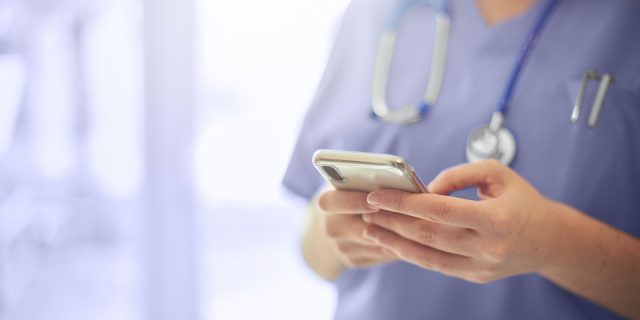Close up of a medical professional's hands holding a phone, wearing scrubs and a stethoscope