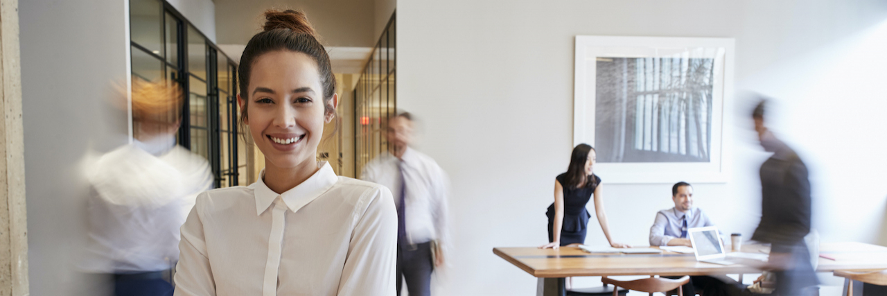 Reflections on Being Openly Autistic in the Workplace Woman smiling with arms crossed at office with people in background blurred behind her