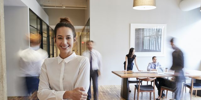Woman smiling with arms crossed at office with people in background blurred behind her