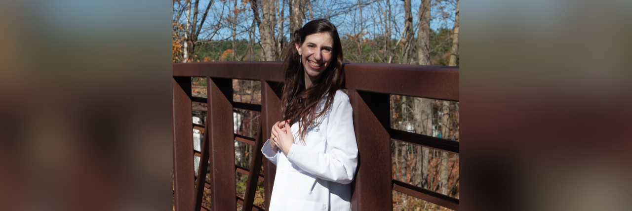 Photo of contributor standing a bridge outside wearing her white doctor's lab coat and smiling