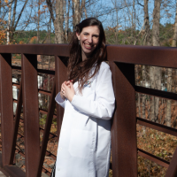Photo of contributor standing a bridge outside wearing her white doctor's lab coat and smiling