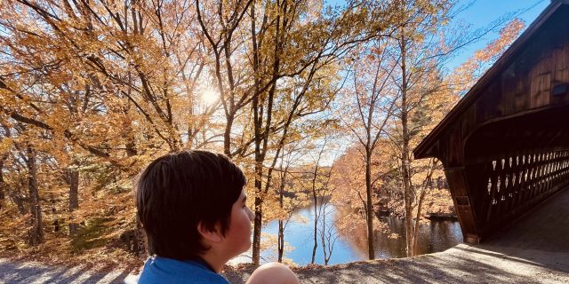 Teetering on the Adolescent Balance Beam With Rare Disease Adolescent sitting near path by a covered bridge and vibrant orange leaves on the trees in the background