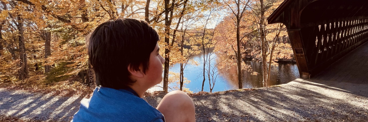Adolescent sitting near path by a covered bridge and vibrant orange leaves on the trees in the background
