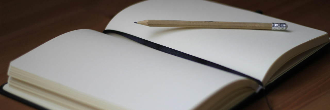 An open blank journal with a black ribbon bookmark down the center with a sharpened pencil on top.