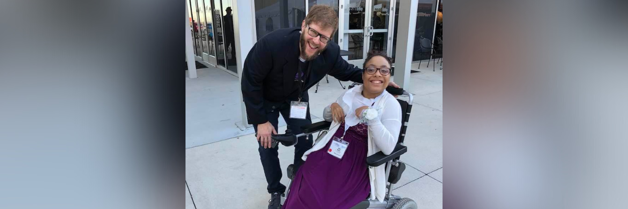 Photo of contributor, a wheelchair user, smiling next to a man who is also smiling and has his arm around the back of her chair