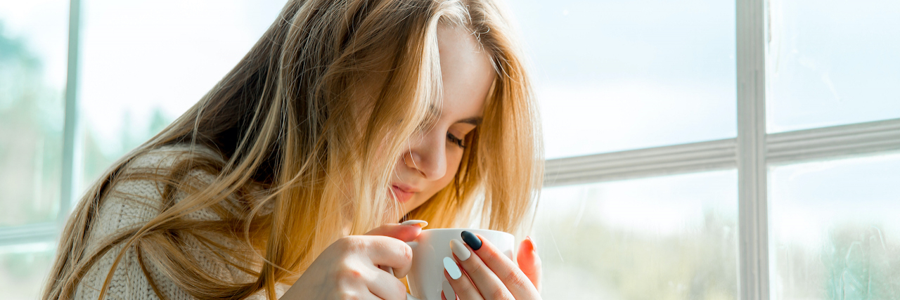 Riding the Roller Coaster of Fluctuating Disability Woman holding a coffee mug sitting next to a window