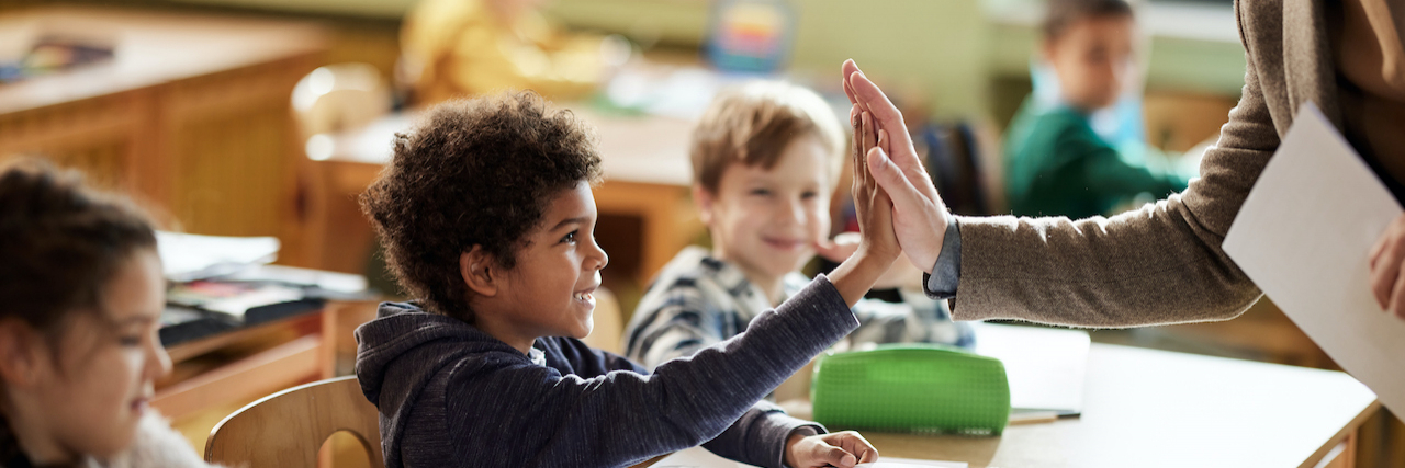 Male teacher giving high-five to a young Black student sitting at a desk class in the classroom.