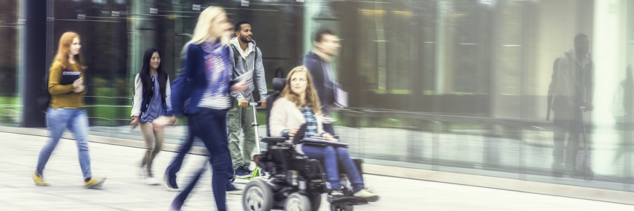Two female friends, one on a wheelchair, walking together in front of a modern university building, other students in backround.