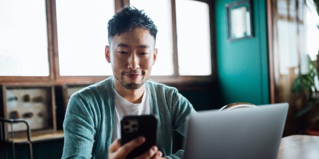 Confident young Asian man looking at smartphone while working on laptop