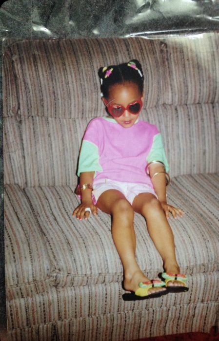 Keisha as a young child, wearing sunglasses and sitting on a couch