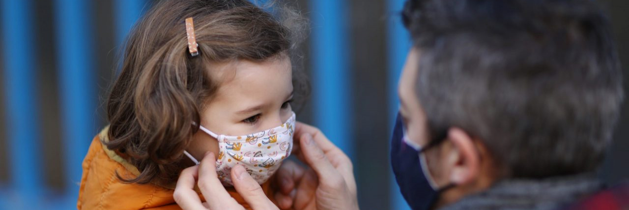 With Cases on the Rise, It's Clear That COVID Isn't Over A father putting a protective face mask on his little daughter in front of school