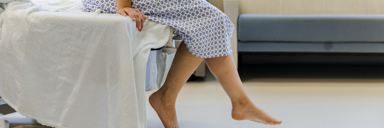 An unrecognizable patient wears a hospital gown and sits on the edge of the hospital bed, dangling their feet.