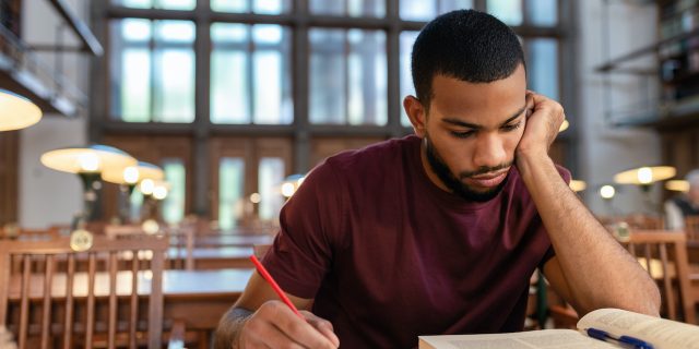 Black man writing in notebook and looking at textbook in library