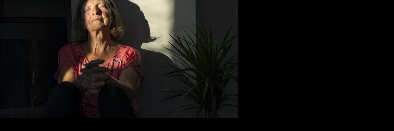 How Chronic Pain and Mental Health Feed Each Other A senior woman in her 70s sits against the wall with the last of the day's sun rays warming her face.