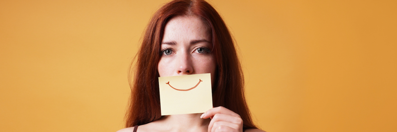 Woman with sad expression in her eyes hiding emotion by covering mouth with fake smile drawn on paper