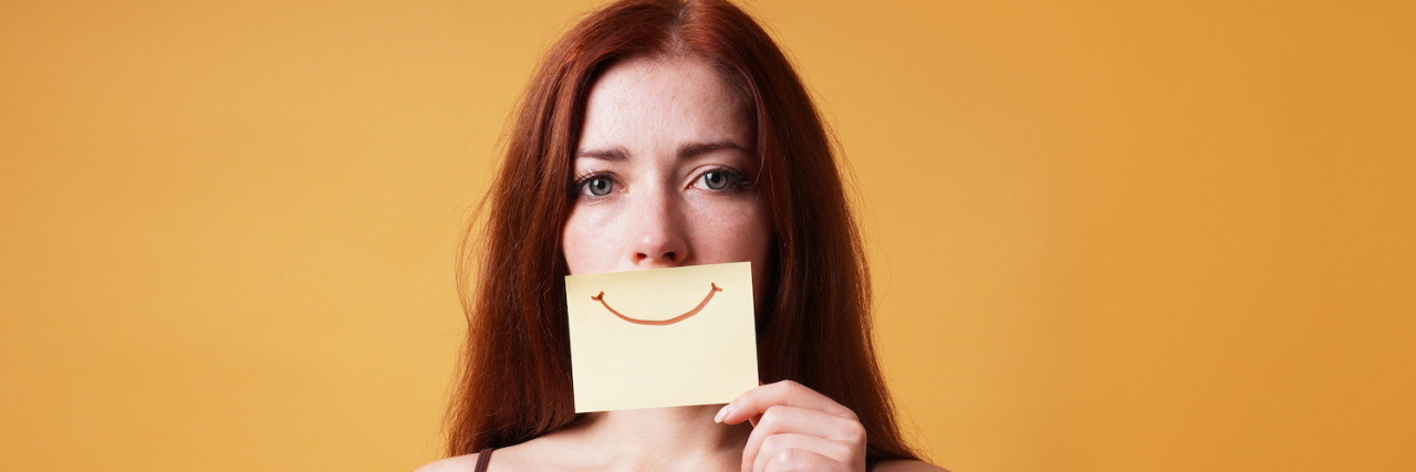 Everything You Need to Know About Smiling Depression Woman with sad expression in her eyes hiding emotion by covering mouth with fake smile drawn on paper