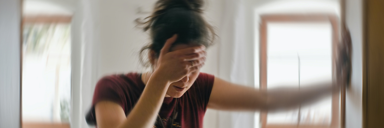 Blurred photo of woman holding her head with one hand and holding herself up against the wall with her other hand, as if experiencing dizziness