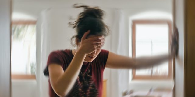 Blurred photo of woman holding her head with one hand and holding herself up against the wall with her other hand, as if experiencing dizziness
