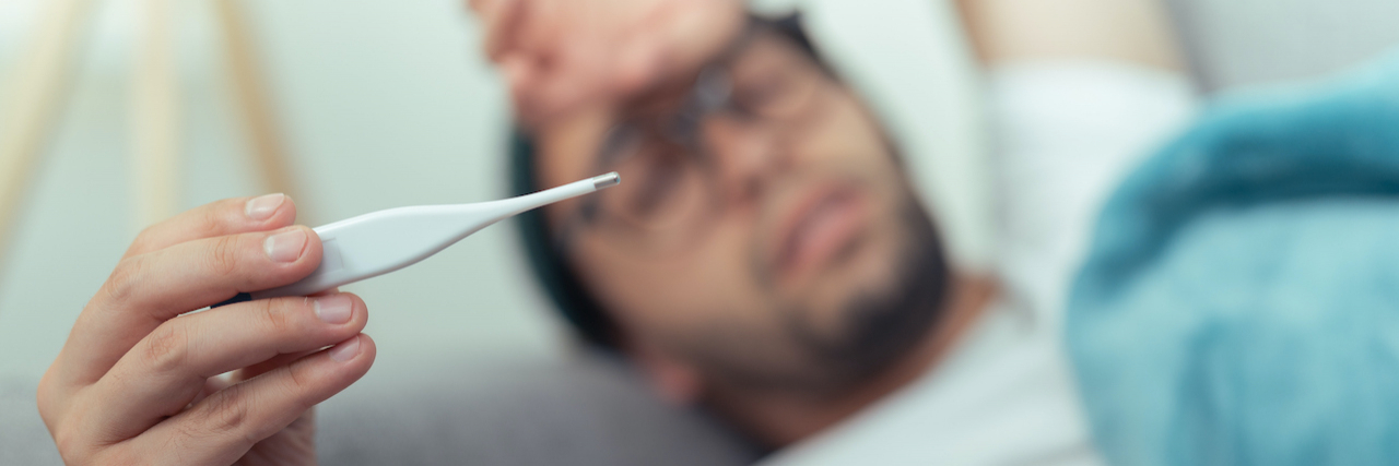 Can a Migraine Cause a Fever? Man laying on couch holding thermometer and his head