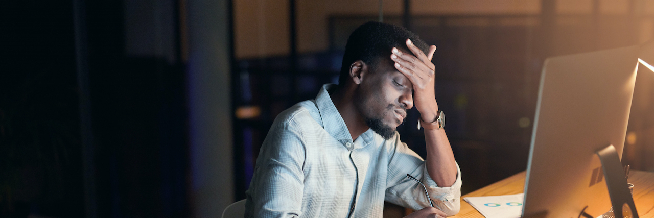 Can Stress Cause Migraine? Black man holding his head with his hand in office in front of computers