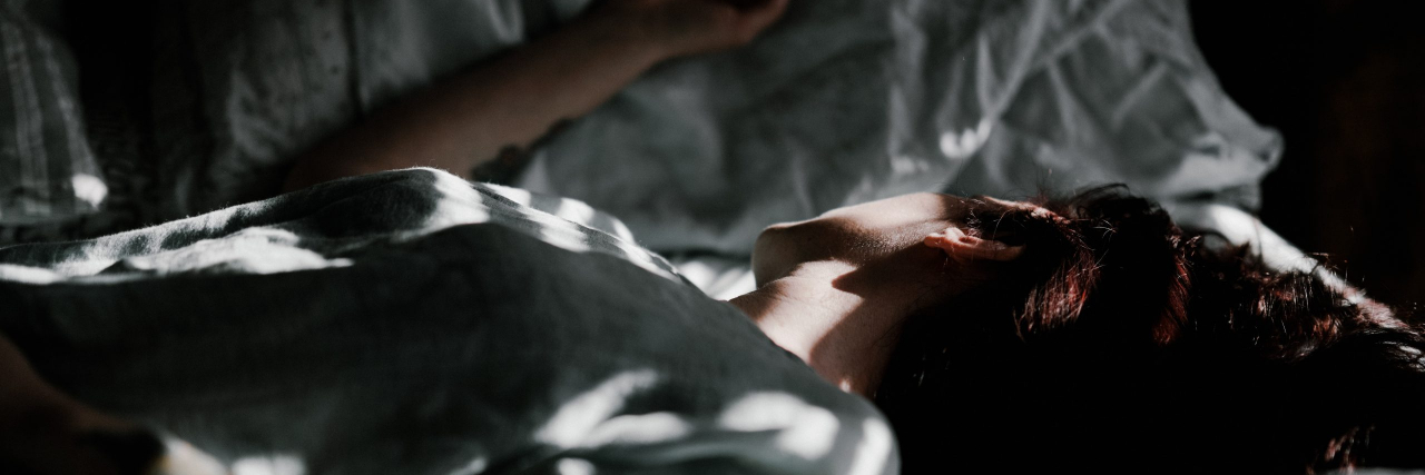 Why Do I Get Sad At Night? A person laying in bed in a dark room with specs of sunlight coming in through blinds.