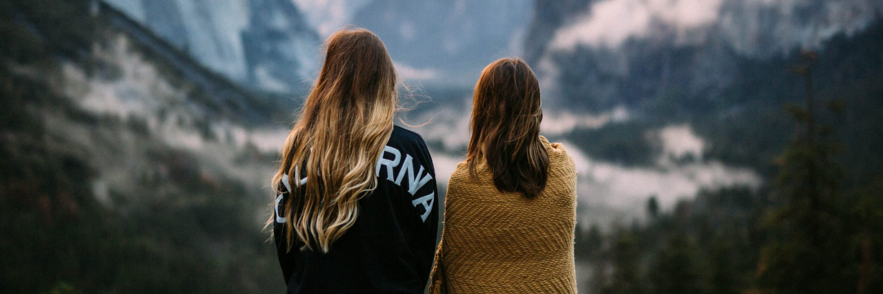 Two women with their backs to the camera looking at out of focused mountain scenery at sunrise.