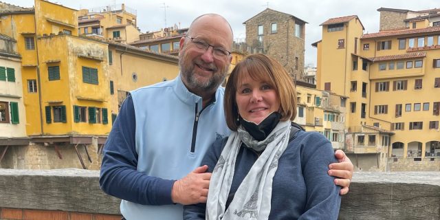 A Musician's Journey to Finding His Rhythm With ALS White man with arms around white woman. Both wear jeans and long sleeves. They stand in front of an orange wall with the view of a European city in the background